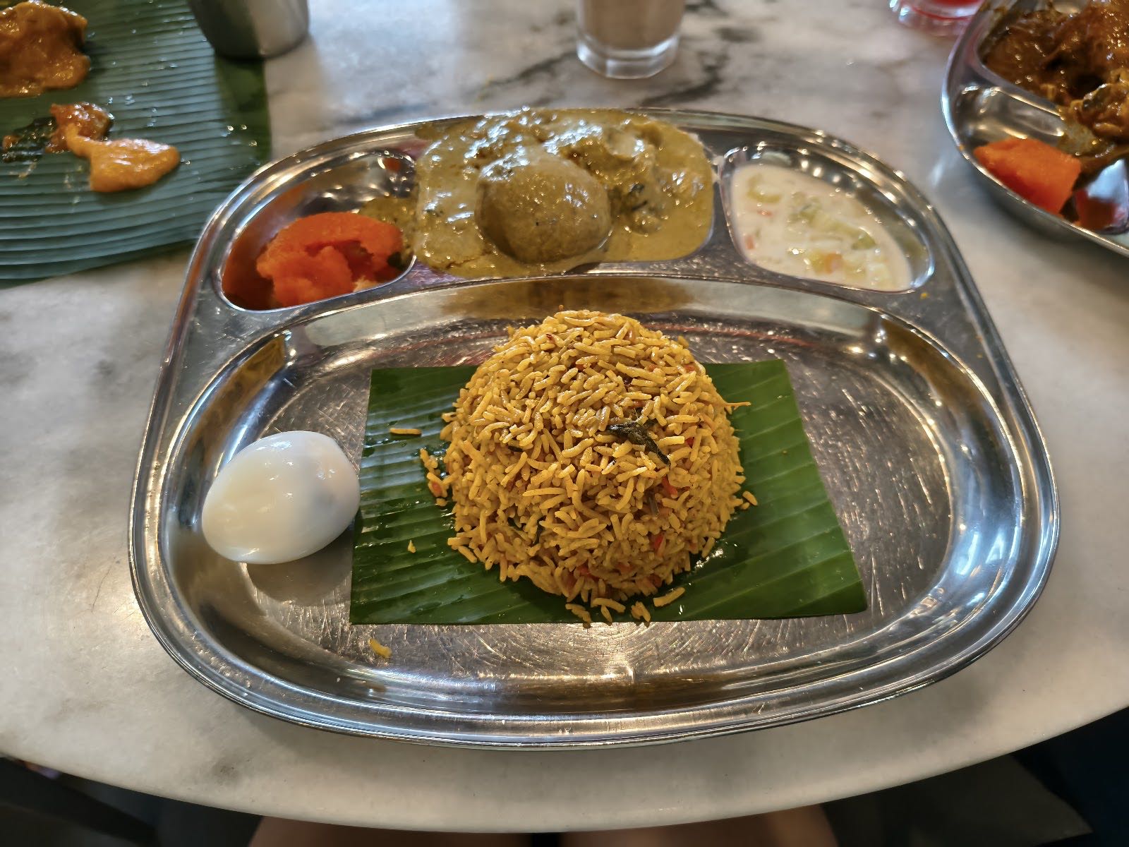 A mound of golden basmati briyani on a banana leaf in a steel thali with raita and a boiled egg