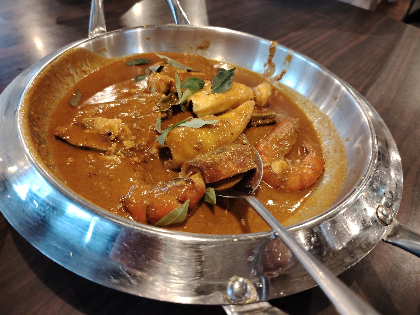 A steel kadai of prawn and seafood curry in a deep orange-brown gravy scattered with curry leaves