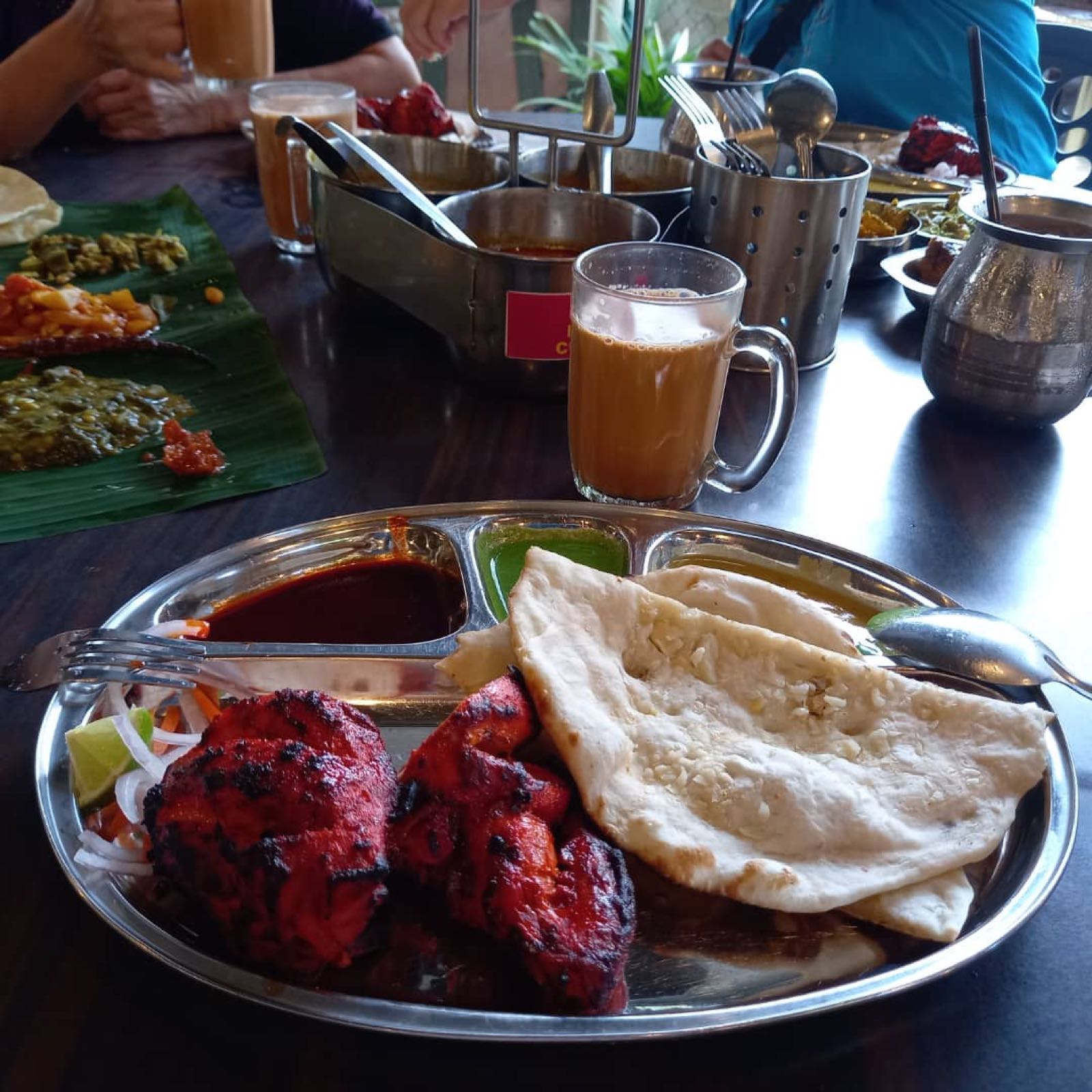 A steel thali of charred tandoori chicken and warm naan beside a glass of teh tarik, with a banana-leaf spread on the communal table behind